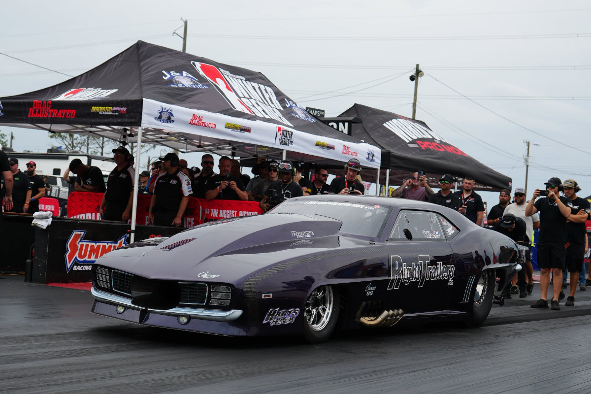 A dark purple drag racing car with custom decals is positioned on a racetrack, with spectators and vendor tents visible in the background under an overcast sky.