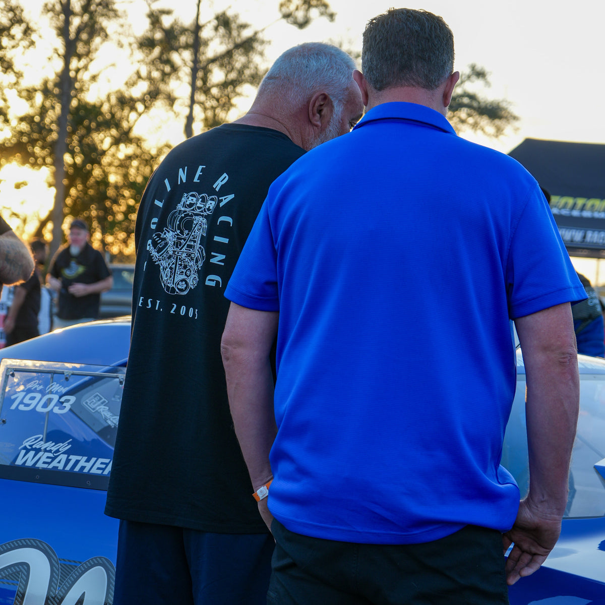 Two men standing next to a blue car with visible branding, trees in the background