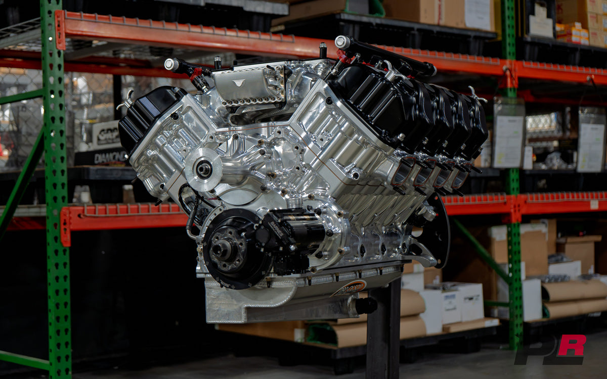 A polished silver and black V8 engine block with a large black intake manifold sits on a stand inside a warehouse or showroom with green and orange shelving in the background.