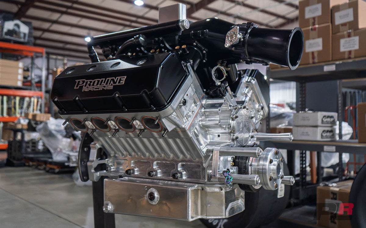 A polished Proline V8 engine block with a black intake manifold and silver aluminum components, displayed on a stand inside a warehouse setting.