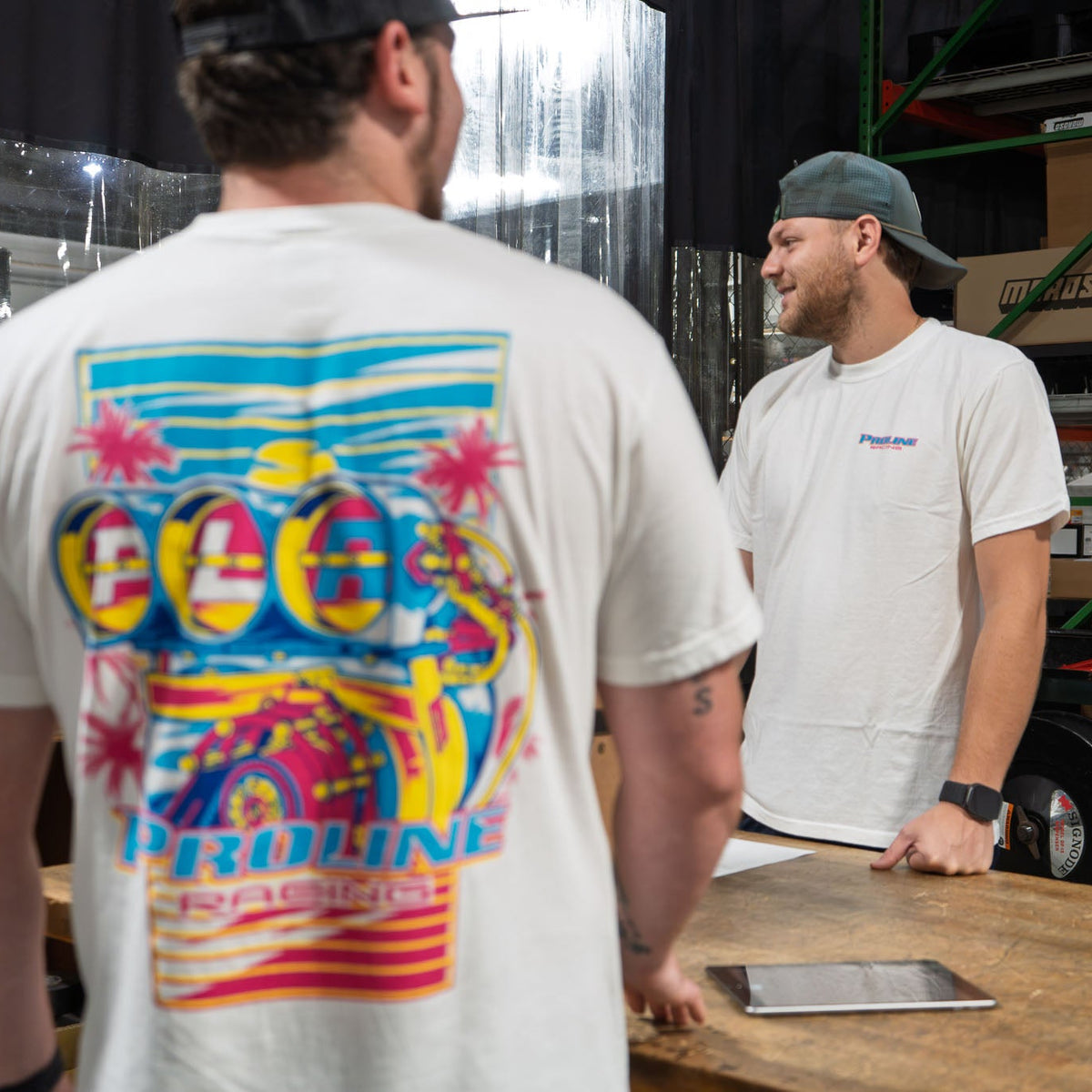 A man wearing a white graphic t-shirt with a colorful engine design stands with his back to the camera, facing another man in a warehouse setting with shelving and boxes in the background.
