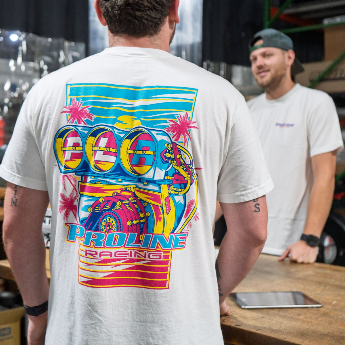 A man wearing a white t-shirt with a large, colorful retro-style graphic on the back, standing in a workshop or garage environment with another person visible in the background.