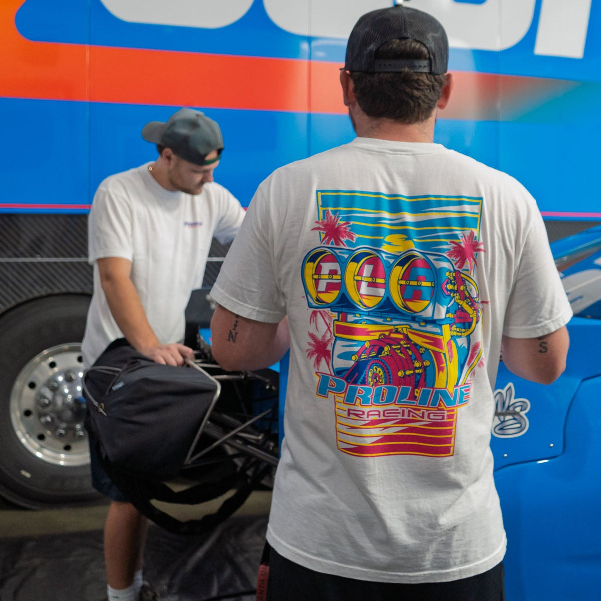 A man wearing a white t-shirt with a colorful Proline Racing Engine Blower graphic on the back stands in front of a blue race truck and blue race car, with another person visible in the background.