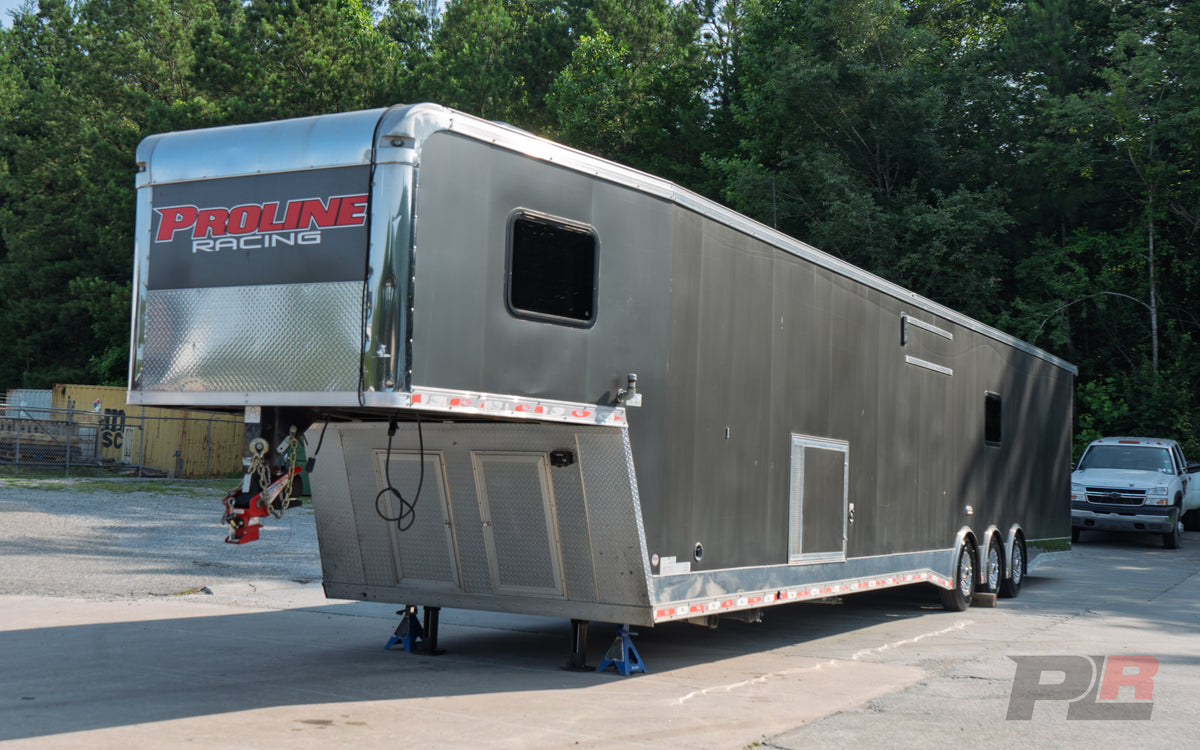 A long silver aluminum race trailer parked outdoors on a paved lot with trees in the background. The front door displays the red and white 'PROLINE RACING' logo, and a white pickup truck is visible on the right side of the frame.